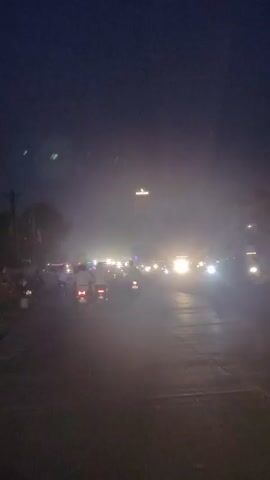 The video captures a nighttime street scene dominated by the dim glow of headlights and streetlights, suggesting a dusky or hazy atmosphere. A procession of motorcycles, ridden by individuals, moves along a paved road. Trees and some structures are visible along the side of the road, partially obscured by the low light and haze. The overall impression is of a busy, albeit dimly lit, urban or semi-urban environment.