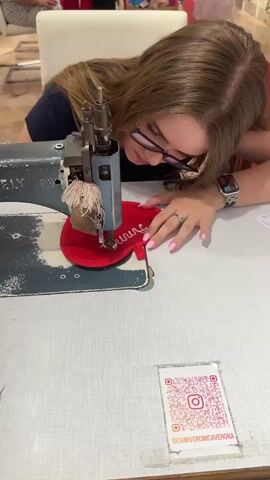 A young woman with long, blonde hair and glasses is leaning over a sewing machine, intently focused on her work. She is wearing a dark blue t-shirt and has her left hand positioned near the needle of the machine, guiding a red, heart-shaped fabric. The fabric has white lettering stitched onto it, appearing to spell out a name or word. A spool of light-colored thread is visible on the sewing machine, and the machine itself is a vintage model, predominantly gray with some visible wear. In the background, out of focus, other people and displays of clothing or fabric can be seen, suggesting a retail or workshop environment. On the table in front of the woman, a QR code is printed on a small white card, displaying a pink Instagram logo and the text "RICAMIVRONICAVVERONA". The overall scene captures a moment of concentration and craftsmanship.