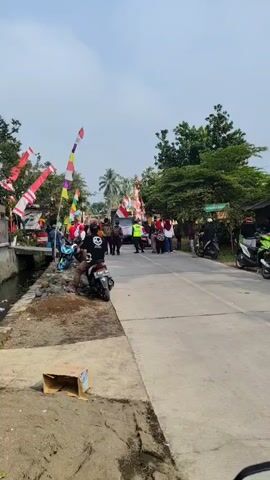 The video shows a street scene with a group of people gathered. Many people are standing on both sides of the road. There are flags and decorations visible, suggesting a celebratory or festive event. Several motorcycles are present, some parked and some being ridden. One person on a motorcycle is in the foreground on the left side of the road, and another is in the mid-ground on the right side of the road. In the distance, more people are visible, and some appear to be interacting. The environment looks like a village or a rural area, with trees and buildings lining the street. The sky is visible and appears to be partly cloudy. There is a small canal or ditch alongside the road on the left. A cardboard box is visible on the ground in the lower left corner.