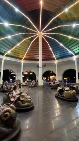 This media file captures a vibrant scene at an amusement park in Nha Trang, Vietnam, likely in the evening given the artificial lighting. The primary activity is a bumper car ride, with several cartoonish, animal-themed bumper cars in motion. The cars, shaped like bulls and mice, are occupied by people of various ages, including children and adults, who appear to be enjoying themselves.

The setting is an enclosed, octagonal structure with a colorful, arched roof adorned with strings of bright lights that radiate outwards from a central point, resembling a starburst. The roof's panels are painted in sections of red, orange, yellow, green, blue, and purple, creating a festive and playful atmosphere. The walls of the structure are white and feature arched openings, through which glimpses of other park attractions and lights can be seen. The floor of the bumper car arena is dark and reflective.

In the foreground, a man in a light-colored shirt and shorts is operating a brown bumper car, his focus on maneuvering the vehicle. Further back, a man in an orange shirt and beige pants is walking across the arena, possibly an attendant overseeing the ride. Other bumper cars are scattered throughout the arena, some occupied by individuals and groups, all participating in the gentle collisions characteristic of a bumper car ride. The overall mood seems cheerful and lively, typical of a fun amusement park experience. No visible text is present in the media.