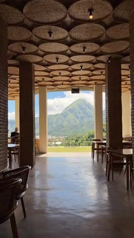 The video shows the interior of a restaurant or cafe with a view of a mountain. The restaurant has a unique ceiling design, with multiple round, woven, straw-like objects arranged in a grid. Support columns are similarly covered. The floor is a polished grey surface reflecting the natural light. Wooden tables and chairs are scattered throughout the space, some occupied by a solitary person. The main focal point is the view through a large opening: a green mountain against a backdrop of blue sky and clouds, with a small town nestled in the foothills.