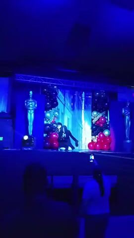 A person is performing on a stage illuminated with blue light. The stage is decorated with two large Oscar-like statues on either side, and balloons in red, black, and silver on the sides of a screen in the center. The screen displays a cityscape with a blue, waterfall-like effect running down. The performer is wearing a black suit and has their arm extended outwards. In the foreground, several people are watching the performance, with one person holding up a phone to record. The environment appears to be a theater or event space.