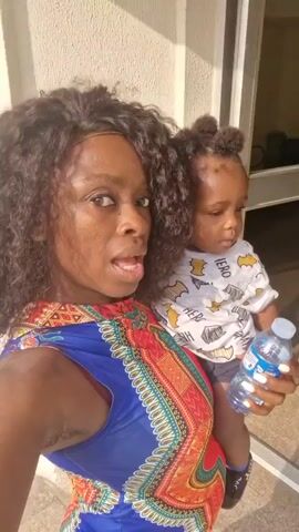 The video shows a woman holding a baby. The woman is smiling and looking at the camera. She has long, curly hair and is wearing a colorful dress. The baby is wearing a white shirt with a superhero pattern. The woman is holding a water bottle in one hand. They are standing in front of a building with a glass door and a sign that says "FLATS RULES AND REGULATIONS." The sign lists rules like "No Smoking cigarettes in the common area."