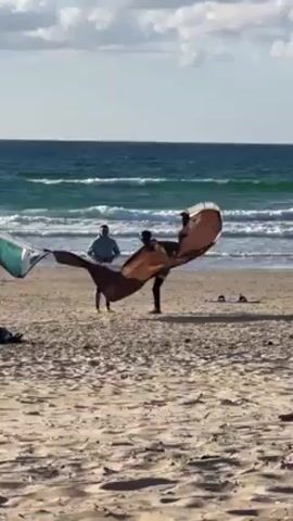 The video shows two people on a sandy beach, preparing a kite. One person is holding the edge of a large, light brown kite, while the other person is working on the body of the kite. A second, turquoise and white kite lies on the ground to their left. In the background, there is a body of water, with small white waves visible. The sky is overcast with some blue sky showing. The sand is marked with various patterns.
