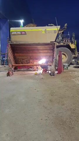 A person is welding inside the bucket of a large yellow construction vehicle. The bucket is open and facing the camera. Sparks are flying from the welding torch. The vehicle has the words "GROUND FORCE 20M" and "CAT" printed on its side. The scene is outdoors at night, with artificial lighting. There are other objects visible in the background, including what appears to be a building and other vehicles.