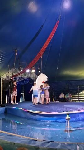 A large, plush white bear costume with black ears and a red bowtie stands on a blue circular stage in a circus tent. Two children, a boy and two girls, stand in front of the bear, facing it. The boy on the left wears a t-shirt and shorts. The girls are dressed in dresses. One girl stands slightly behind the bear, holding a music stand. The bear's paws are raised, and it appears to be interacting with the children. Another boy stands to the far left of the stage, wearing a red shirt and shorts, and appears to be watching the performance. A red fabric hangs from the ceiling of the tent. The stage is surrounded by a shallow pool of water. Lights are visible overhead, and there are curtains and decorations in the background.
