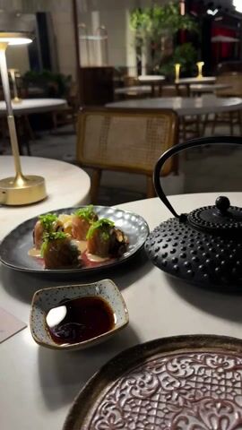 The video showcases a neatly arranged outdoor dining table at a restaurant during nighttime. The table is set with a plate of sushi rolls drizzled with a light sauce and garnished with green herbs, a small dish filled with dark soy sauce, and a decorative black teapot. A menu card with a QR code is also present. The table is illuminated by a small, golden-colored table lamp. In the background, several other tables and chairs are visible, indicating an open-air restaurant setting. The lighting is subdued, creating a relaxed and intimate ambiance.