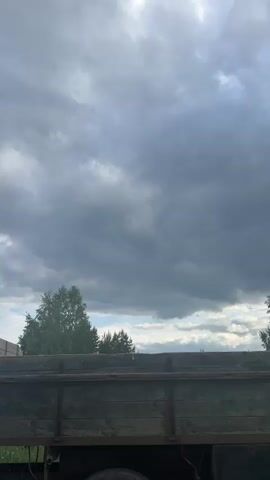 The video shows a landscape with a cloudy sky overhead. The camera pans from left to right, revealing a wooden truck bed in the foreground, followed by trees and a field in the background. A small pile of dirt is also visible in the field. The sky is filled with dark, gray clouds, but there are also patches of blue sky and sunlight peeking through.