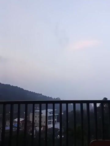 The video pans across a wooden table on a balcony overlooking a landscape of trees and buildings in a valley, possibly in the early morning or late evening as the sky is overcast and dimly lit. The table has two cups filled with a dark liquid, possibly coffee, an ashtray filled with cigarette butts and tobacco, a lighter, and a pack of cigarettes. The background is partially obscured by a black railing.
