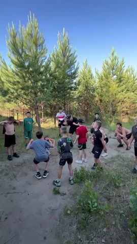 A group of young boys and one adult male are gathered in a clearing in a forest. The adult male appears to be instructing or leading the boys in some form of exercise or activity. The boys are positioned in a rough circle, and many of them are in a squatting or crouched position. They are all dressed in casual clothing. The environment is natural, with pine trees and undergrowth visible. The sky is clear and blue, suggesting it is daytime and likely sunny. The ground is dirt and covered with some scattered leaves and twigs. The overall impression is of an outdoor training session or game.