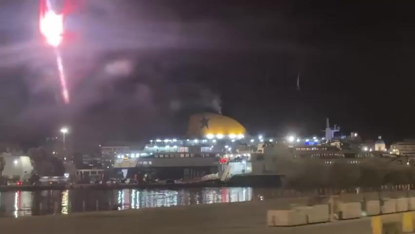 The video shows a night scene at a harbor. A ferry with a prominent yellow funnel and blue star is docked. Lights illuminate the ferry and the surrounding area. In the sky, there are streaks of red and white light, resembling fireworks or flares. The camera pans slightly to the right, revealing a road with yellow barriers and a directional road sign. The water reflects the lights of the harbor, creating a shimmering effect. The overall atmosphere is dark, but the lights add a sense of activity to the scene.