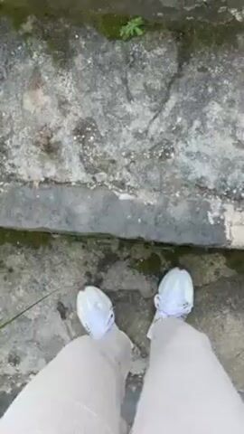A person is walking up a set of stone steps. They are wearing light tan ribbed pants and white tennis shoes with light blue accents. The steps are weathered and covered in lichen and moss in some areas. The camera is positioned directly above the person, providing a first-person perspective of their ascent. The focus remains on their legs and feet as they move upwards.