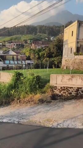 The video shows a view of a rural mountainside village. The camera pans across a road with houses and vegetation on either side. In the background, a mountain looms under a cloudy sky. The footage appears to be shot from a moving vehicle. There are no people visible in the video.