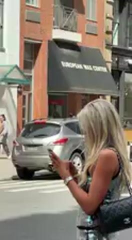 The video captures a woman with long, light blonde hair, standing on what appears to be a city sidewalk, engrossed in her smartphone. She is wearing a silver-toned, sparkly, sleeveless top and a matching skirt or shorts, with dark bra straps visible. A black quilted shoulder bag, consistent with the style of a Chanel classic flap bag, hangs from her right shoulder. She holds her phone in both hands, looking down at the screen.

In the background, an urban street is visible with white crosswalk lines. A silver SUV passes slowly, and other vehicles can be inferred. Buildings with brick facades line the street. One prominent building has a dark awning over a storefront, clearly displaying the text "EUROPEAN WAX CENTER." A blue public recycling or trash bin, typical of New York City, stands near the curb.

The scene is brightly lit by natural sunlight, suggesting a clear day, likely during the daytime. A wooden barrier or railing is partially visible in the immediate foreground, obscuring the lower part of the scene. The overall setting strongly suggests a bustling street in New York, United States.