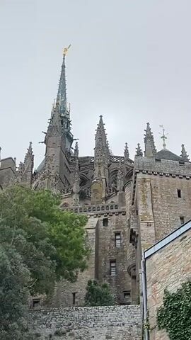 The video features a low-angle shot of a large, ancient, grey stone architectural complex, likely a cathedral or abbey, against an overcast sky. A prominent, slender central spire with a golden finial dominates the upper structure, flanked by numerous smaller spires and gothic architectural details. The camera slowly pans downwards, revealing the complex is built on an elevated position, with multiple levels of stone walls, terraces, and smaller buildings. Lush green vegetation, including trees, bushes, and ivy climbing the walls, is visible in the foreground and mid-ground.