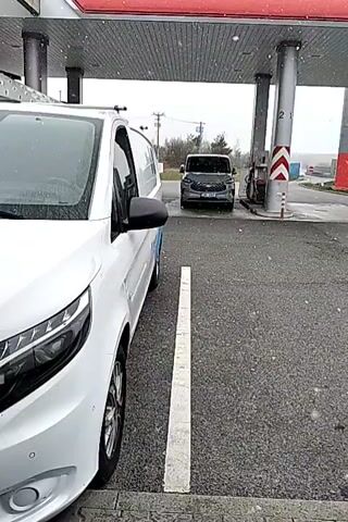 The video shows a white van parked at a gas station, with another gray van parked at a gas pump in front of it. The gas station is covered, with columns supporting the roof. The weather appears to be rainy or snowy, with visible precipitation. The background shows telephone poles and a distant landscape.