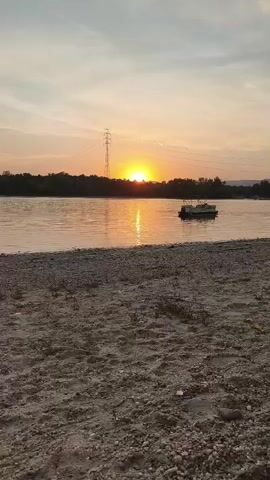 This media file captures a serene sunset over a wide river, likely the Danube, with Dunakeszi, Hungary, as the backdrop. The foreground features a sandy and pebbly shore, creating a natural beach. In the tranquil water, a pontoon boat is visible, suggesting a leisurely atmosphere. Across the river, a treeline defines the horizon, broken by a prominent electricity pylon. The sky is painted with the soft hues of sunset – warm oranges and yellows reflecting on the water, transitioning into a pale gray and blue at the top of the frame. A few wispy clouds are scattered across the sky. The overall mood is peaceful and calm, evoking a sense of tranquility. The time of day is clearly sunset, as indicated by the low position of the sun and the warm colors in the sky and water. The location cues, such as the pylon and the characteristic landscape, align with the provided context of Dunakeszi, Hungary. There is no visible text in this media file.