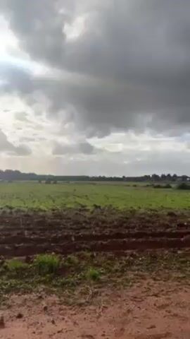 The video shows a partially plowed field under a sky full of clouds.  The sun is shining brightly behind the clouds, creating a glare. Part of the field is covered with green vegetation. A building can be seen in the distance. The foreground consists of dirt. The camera pans slowly upward.