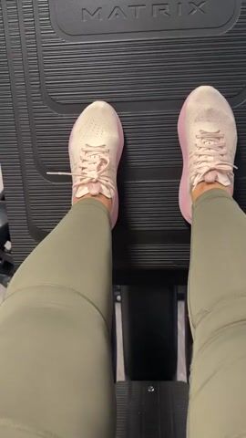 A first-person, high-angle shot shows a person's feet and lower legs as they use a step mill or stair climber machine. The person is wearing light olive green or khaki-colored leggings and light pink athletic shoes with white laces and soles. The feet are positioned on the black, ribbed steps of the exercise machine, moving rhythmically up and down as if climbing stairs. The brand name "MATRIX" is clearly visible in white lettering on the black top surface of the machine, directly in front of the person's feet. The setting appears to be a gym or fitness center.