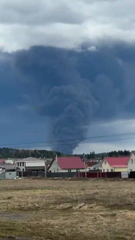 The video captures a large, dark grey to black smoke plume rising dramatically into an overcast sky. The massive column of smoke billows upwards, expanding widely at its peak, dominating the distant horizon. Below the towering smoke, a rural or semi-rural landscape unfolds, featuring several detached houses with varying roof colors (red, brown, and light-colored) scattered across a sparse, earthy field. Power lines and utility poles are visible in the foreground and mid-ground. A dense line of trees, likely a forest, stretches across the background, situated beneath the origin point of the smoke. The sky above is heavily clouded with dark, somber tones, enhancing the ominous appearance of the smoke. The scene depicts a significant incident, possibly a large fire, in the vicinity of residential buildings in Kosoturikha, Russia, under daytime conditions.