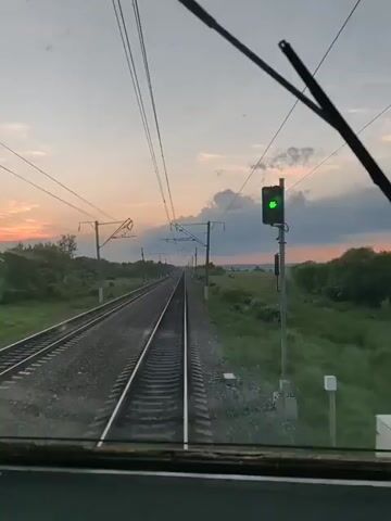 The video captures a first-person perspective from inside a train cab, looking out through the front window. The camera is positioned to show the tracks stretching out ahead, leading towards a horizon painted with the soft hues of sunset. The sky is a gradient of pale blue, transitioning to pink and orange, with scattered clouds tinged by the fading light.

The train is traveling along a double set of railway tracks, flanked by lush green fields and trees on either side. Overhead, a complex network of power lines and support poles stretches into the distance, indicating an electrified railway line.

The time of day is clearly dusk, as evidenced by the sunset colors and the darkening landscape. The weather appears to be mild, with no rain or harsh conditions visible. The overall mood is one of peaceful travel, with the rhythmic movement of the train and the scenic, tranquil countryside creating a sense of calm.

There is some visible text in the lower left corner of the frame, displaying the number "68" on a digital display. This could potentially be a speed indicator or another form of data relevant to the train's operation. The location context provided states Arkhara, Russia, which helps to place the scene geographically. The video appears to be a continuous shot, with no discernible beginning or end point of an event beyond the ongoing journey.