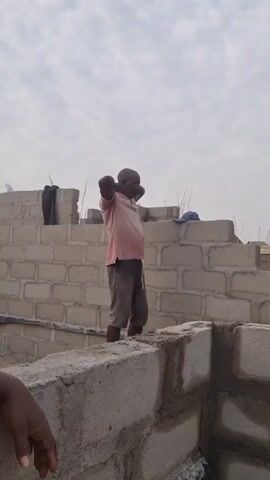 The video shows a man standing on a partially constructed brick wall. He is wearing a pink short-sleeved shirt and grey shorts. The wall appears to be part of a building under construction, made of light grey cinder blocks. The sky is overcast. A small section of another person's arm is visible in the lower left corner of the frame. The man stretches his arms over his head, then puts his hands behind his head and looks around. A blue hat and a dark bag can be seen on the brick wall behind him.