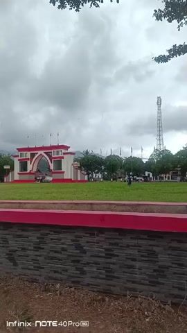 The video begins with a wide shot of an outdoor scene under a cloudy sky. In the foreground, there is a low stone wall with a red border. Beyond the wall is a grassy area with a red border. In the midground, there is a large, arch-shaped structure painted white and red, with flags on top. Several cars are parked near this structure. Further back, there are more trees, a tall communication tower, and some distant hills. A few people can be seen walking around in the midground and background. The camera pans slowly to the right, revealing more of the open field and some buildings. The video concludes with a shot of paved ground and more grassy areas with red-bordered planters.