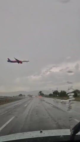 The video shows an airplane flying low in the sky. The perspective is from inside a vehicle, looking out through the windshield, which is wet with rain. The road ahead is wet and reflecting the lights of other vehicles, suggesting it is nighttime or dusk. The sky is overcast and gray, indicating rainy weather. Trees and hills are visible in the distance. The airplane appears to be a commercial airliner with a pink and white livery. It is flying at a low altitude, likely on its approach to an airport. There is no visible text in the video. The overall mood is somewhat dramatic due to the low-flying plane and the stormy weather.