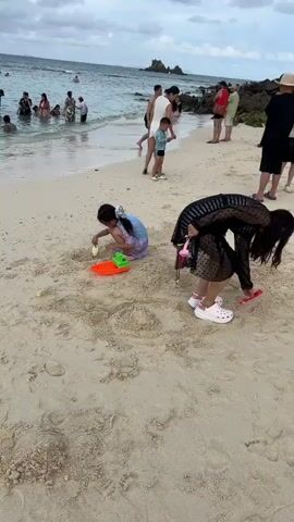 The video captures a beach scene. In the foreground, two females are on the sand, one a young girl playing with sand toys, and the other an adult woman bending down, holding what appears to be a red object. In the background, several people are visible near the water, some wading in the ocean. The environment includes a sandy beach, the ocean, and a cloudy sky.