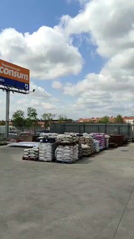 The camera pans across a concrete lot behind a supermarket, under a partially cloudy blue sky.  In the foreground are stacks of bagged goods, arranged in rows on pallets.  Behind them is a metal fence, then the buildings of a small town.  A DACHSER truck, with a yellow and blue paint scheme, drives into the shot and stops in the lot.