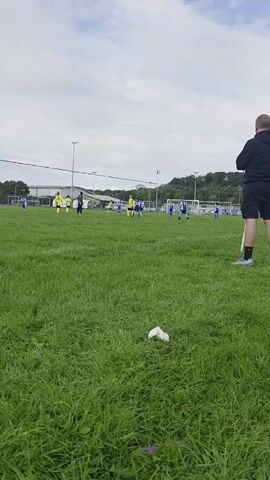 The video opens with a close-up of a person holding a yellow dandelion in their hand, with the stem blurred. The background is a grassy field where a soccer game is in progress. Several young players, mostly wearing yellow jerseys and blue shorts, are scattered across the field. A few adults, likely coaches or referees, are also present. The sky is overcast with scattered clouds. In the distance, there are buildings and trees. The camera is positioned low to the ground, looking up at the field. The video pans slightly to the left, showing more of the soccer game. There is some trash, a white piece of paper or plastic, on the grass in the foreground. The overall impression is a casual outdoor sports event.