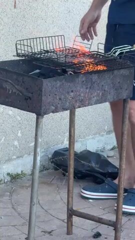 A man is shown grilling outdoors. He is wearing a dark blue short-sleeved button-down shirt and matching dark blue shorts, along with blue Crocs. The man is actively tending to a barbecue grill, which is in operation and has flames visible. He is interacting with the grill, seemingly adjusting the grate or food. In the background, there is a building with a neutral-colored wall and a window with white blinds. The ground appears to be paved with interlocking bricks. A black plastic bag is visible on the ground near the grill. There is also a metal bowl containing food items on the ground to the right of the grill. The overall environment suggests a casual outdoor setting, possibly a patio or backyard.