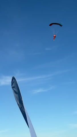 A skydiver wearing an orange jumpsuit and helmet descends with a red and blue parachute against a clear blue sky. The skydiver is in mid-air, approaching a green field below. The parachute is open and appears to be stable. The perspective of the camera is from below the skydiver, looking up.