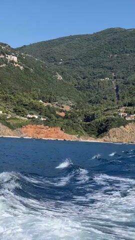 The video shows a view from a boat moving through blue water. The camera angle is low and tilted slightly upwards. In the foreground, white frothy wake churns behind the boat. The water is a deep blue, with some choppy waves and whitecaps visible. In the middle ground, a coastline with steep, green, tree-covered hills slopes down to the sea. There are small white buildings scattered on the hillsides. A section of the coastline appears to be a reddish-brown cliff face. The sky is a clear, bright blue.