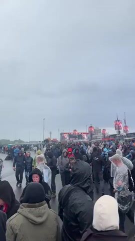 A large group of people are walking on a paved surface during what appears to be a rainy day. Many are wearing rain ponchos and jackets. In the distance, there is a large stage with bright lights. Several tall structures with banners are visible as well. The sky is overcast and grey. Some individuals are looking at their phones. The overall impression is a crowd attending an outdoor event in inclement weather.