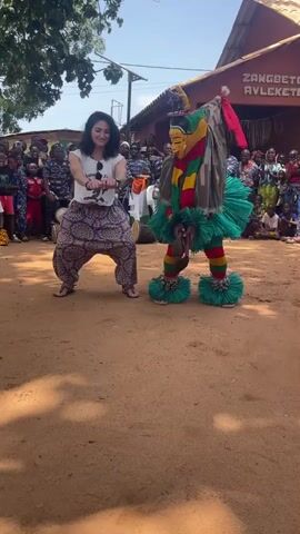 The video captures a vibrant outdoor scene likely in West Africa, possibly Benin or Togo, given the signage in what appears to be Fon language. In the foreground, a young woman with dark curly hair, wearing a white t-shirt and loose patterned pants, performs a dance. She is positioned to the left of the frame, facing slightly right, with her arms bent and hands held out in front of her. To her right, a masked performer, or Zangbeto, dominates the center of the frame. The Zangbeto is elaborately costumed, featuring a tall, pointed mask painted with yellow, red, and green. Its body is adorned with a striped fabric, a green ruffled skirt, and red leggings, all accentuated by large, green, fluffy ankle decorations. The masked figure is in a low, crouched dance posture. The interaction between the woman and the Zangbeto suggests a performance or cultural exchange. Behind them, a crowd of onlookers, predominantly children and adults in casual attire, are gathered, creating a sense of community and shared experience. The background reveals a building with a sign that reads "ZANGBETOR AVLEKETOU," and trees with lush green foliage, indicating an open, possibly rural or village setting under a bright, sunny sky. The ground is composed of reddish-brown earth, common in tropical regions. The overall atmosphere is lively and festive.