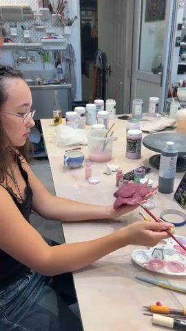 A young woman with glasses and dark curly hair is sitting at a table in a workshop. She is wearing a black top with lace trim and blue jeans. She is holding a piece of reddish-brown clay in her left hand and is painting it with a brush from a palette with pink and white paint. The table is covered with various pottery supplies, including brushes, jars of colored glaze, bowls, cups, and tools. In the background, shelves are filled with more supplies and finished pottery pieces. The workshop appears to be well-lit and organized, with a window showing greenery outside. The scene suggests a creative and focused atmosphere.