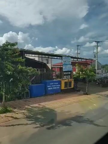The video pans across a street with buildings and vehicles. On the left, there are two yellow generators. Further down the street, a white pickup truck is parked in front of a building. A blue truck with a tarp-covered bed is driving down the street. In the background, there are power lines and trees. The sky is cloudy. The street appears to be wet, suggesting it may have rained recently.