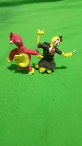 The video shows two clay figures on a green surface. The figure on the left is red with yellow accents, has two large white eyes, and appears to be lunging forward. The figure on the right is yellow and black, wearing a black outfit with yellow trim and a black hat. It has black stripes across its face and appears to be reaching out with its left arm. The background is a solid green surface, likely fabric.