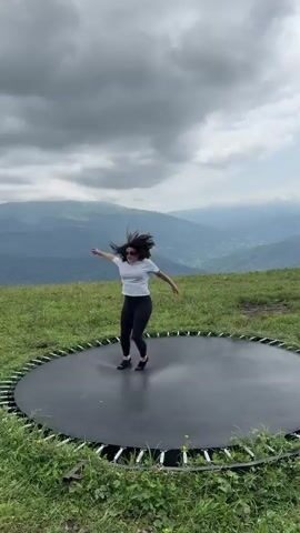 A young woman is jumping on a trampoline outdoors on a grassy hill. She is wearing a grey t-shirt and black leggings. The trampoline is round and black. In the background, there are mountains and a cloudy sky. The woman is in mid-air, jumping, and then she spins around.