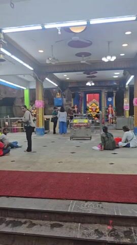 The video shows the inside of a temple located inside a cave. The temple area is well-lit with artificial lights and has pillars supporting the structure. Some people are present, including individuals in casual clothing and one man wearing a traditional red garment with his chest bare. A red carpet is laid out in part of the temple.  The camera then pans upwards to showcase the natural rock walls of the cave, and continues panning up to reveal the opening at the top of the cave, allowing sunlight to filter through. Green vegetation grows along the edges of the cave opening.