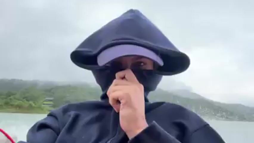 The video shows two women on a boat. One woman is wearing a dark blue hooded jacket and a purple baseball cap with a logo. She has the hood up and the jacket zipped up to cover part of her face. The other woman is next to her and is wearing a gray hooded jacket. They are on a lake or river, and the boat is moving quickly. There are trees and hills visible in the background. The sky is overcast.