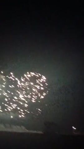 The video shows a nighttime sky filled with bright white fireworks exploding in a cluster. The bottom of the frame shows a dark landscape with a few faint lights, possibly from distant buildings or streetlights. The fireworks are the main focus, creating a dazzling display against the dark sky.