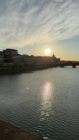 The video shows a sunset over a river in an Italian city, presumably Florence. The camera pans from left to right across the water, revealing the reflection of the sun, the city skyline including a dome, a bridge, and buildings along the riverbank. People can be seen sitting and standing along the stone wall that runs along the river. The video ends focused on the buildings and people.