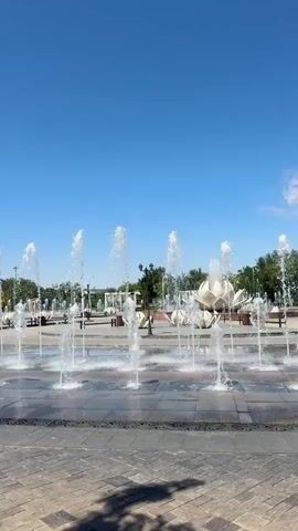 A wide shot captures a public square with multiple water fountains spraying into the air under a clear blue sky. The fountains are arranged in rows, creating a dynamic visual effect as water jets shoot upwards and fall back down. In the background, there are trees, benches, and some decorative structures that appear to be shaped like large flowers. The ground is paved with cobblestones and stone tiles. The scene is well-lit, suggesting it is daytime.