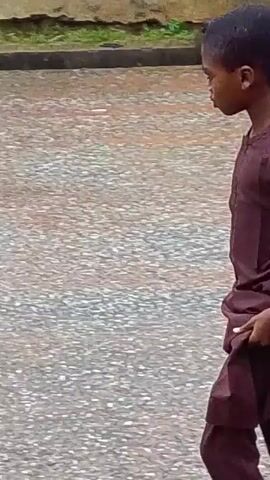 The video shows a young boy walking on a wet, paved surface. He is wearing a long, loose-fitting dark colored garment. In the first few seconds, he is seen from the side, walking away from the camera and reaching into his garment. The scene then shifts to show the boy from a slightly different angle, still walking away and seeming to be touching his face. In the background, there are buildings with corrugated metal roofs and some vegetation. The ground appears to be a road or pathway, and there are puddles of water, suggesting it may have recently rained. The boy is the sole focus of the video, and there are no other people clearly visible interacting with him.