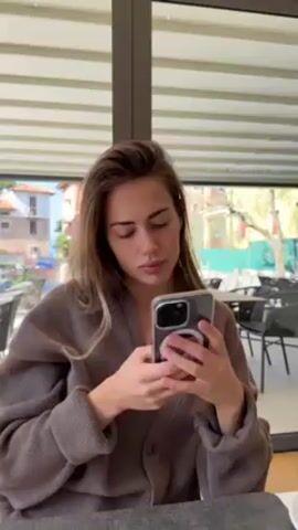 A young woman with long blonde-brown hair is sitting at a table and looking at her phone. She is wearing a brown cardigan. The phone has a clear case and a circular attachment on the back. Through the large window behind her, there is a view of buildings and trees. There are tables and chairs visible both inside the room where she's sitting and outside. The window has white horizontal blinds at the top.