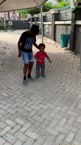 The video shows two children walking on a herringbone-patterned brick driveway. The elder child, a boy, is wearing a black t-shirt with white lettering, jean shorts, grey shoes and black socks. He has a dark complexion and short black hair. He holds the hand of a younger child, also a boy, who wears a red t-shirt with stars, grey pants, and green shoes. This younger child has dark complexion and short curly black hair. The older boy kicks a plastic water bottle on the ground. Behind them is a grey fence and a partially visible structure that looks like a carport. A green trash can is visible near the fence.