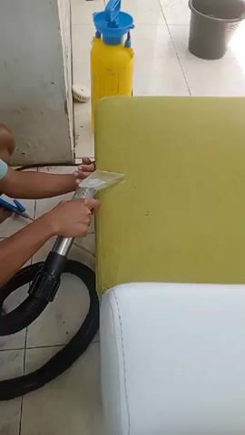 A person kneels on a tiled floor next to a sofa. The sofa has a yellow section and a white section. The person is holding a cleaning tool with a hose attached to it. The tool is being used to clean the white section of the sofa. A yellow spray bottle and a dark bucket are visible in the background. The person is wearing a blue t-shirt.