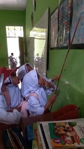 A vertical medium shot shows a group of young students, likely elementary school age, in a classroom setting. The students are wearing white uniforms with headscarves, suggesting they attend a religious school. Three students are prominently featured in the foreground, looking towards a chart on the green wall. One student on the right, wearing a white headscarf and uniform, points with a long stick towards the chart. Another student to her left, also in a white headscarf and uniform, mimics the pointing gesture. A third student, partially visible on the far left, wears a red cap and a white uniform. Their attention is directed at a chart on the wall which displays letters of the alphabet and numbers in a grid format, possibly a learning aid for early literacy. The classroom appears to be simple, with a green wall and a glimpse of a doorway with sunlight streaming in, indicating daylight hours. Another student, further back and out of focus, is also visible near the doorway. The students are engaged in a learning activity, with the teacher or a student leader using the stick to guide their focus on the educational chart. The overall impression is one of a typical classroom scene focused on instruction.